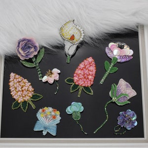 Könnte beinhalten: Sortiment an bunten Perlenblumenbroschen, darunter eine rosa und weiße Blume, eine lila Blume, eine weiße Calla, eine rosa Erdbeere, eine blaue Blume, eine lila Blume und ein Blumenstrauß.