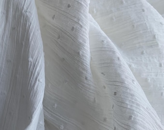 May include: Close-up of white fabric with a textured, dotted pattern. The fabric is draped, showing folds and shadows that highlight the texture. The material appears soft and lightweight, suitable for clothing or home decor.