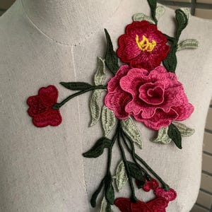 May include: Embroidered floral applique featuring a cluster of red and pink flowers with green leaves. The flowers have detailed stitching and a textured appearance. The applique is likely for clothing or craft projects.