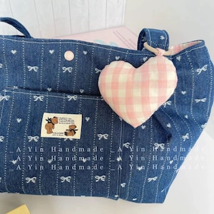 Puede incluir: Una bolsa de mano de lona azul con corazones y lazos blancos. La bolsa tiene un dije en forma de coraz&oacute;n de cuadros rosa y blanco sujeto al asa. La bolsa tiene un bolsillo en la parte delantera con un logotipo de oso marr&oacute;n y el texto "AYin Handmade".