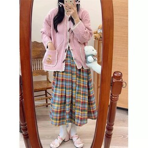 K&ouml;nnte beinhalten: Rosa Zopfstrick-Cardigan mit Holzkn&ouml;pfen und einer Tasche mit einem gestickten B&auml;ren. Kombiniert mit einem karierten Rock in Blau, Gelb und Rot und rosa Schuhen. Das Outfit spiegelt sich in einem Holzrahmen wider.