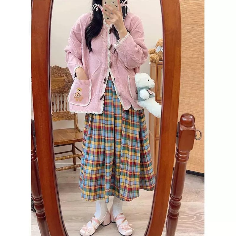 K&ouml;nnte beinhalten: Rosa Zopfstrick-Cardigan mit Holzkn&ouml;pfen und einer Tasche mit einem gestickten B&auml;ren. Kombiniert mit einem karierten Rock in Blau, Gelb und Rot und rosa Schuhen. Das Outfit spiegelt sich in einem Holzrahmen wider.