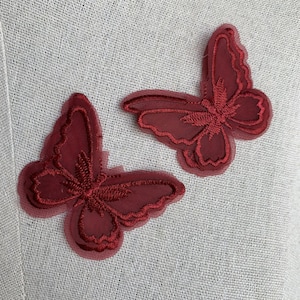 May include: Two maroon butterfly appliques. Each butterfly is intricately embroidered with a detailed wing pattern. The appliques are made of a sheer fabric, giving them a delicate appearance. They are set against a light-coloured, textured background.
