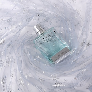 May include: A clear glass bottle of cologne with a blue liquid. The bottle has a silver label with the text "LOVALI ESSENTIAL MEN" and "EAU DE COLOGNE 1.00 FL. OZ. 100ML. E". The bottle is sitting on a white tulle fabric with silver glitter snowflakes.