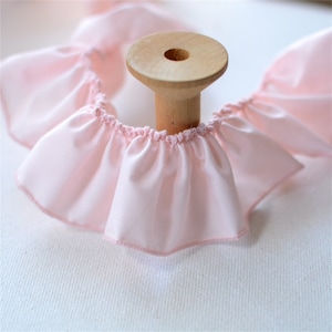 May include: A spool of light pink ruffled fabric. The fabric is gathered and sewn to create a ruffled edge. The spool is made of light-colored wood. The fabric is a soft, pastel pink color.