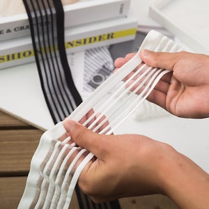 Könnte beinhalten: Eine Person hält ein weißes, durchsichtiges Gummiband mit vertikalen Streifen. Das Band ist teilweise gedehnt und zeigt seine Textur. Ein schwarzes Band mit ähnlichen Streifen ist im Hintergrund zu sehen, zusammen mit Büchern.