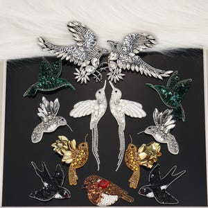 Op de afbeelding: Een collectie vogelvormige geborduurde patches met diverse ontwerpen. Inbegrepen zijn zilveren paillettenvogels, groene kristallen kolibries, witte kralenvogels met lange staartfranjes, gouden paillettenvogels en een bruine vogel met een rode kraal.