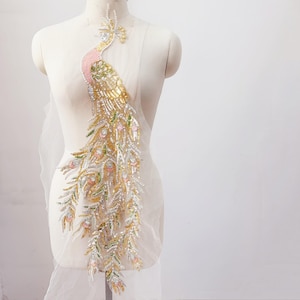 May include: A decorative peacock applique with a pink, gold, and green color scheme. The applique is embellished with sequins and beads, and is attached to a sheer, white fabric backing. The peacock design is displayed on a mannequin.