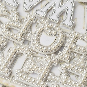 May include: A close-up of a collection of white felt letters decorated with pearl beads. The letters are arranged in a random order and are all the same size.