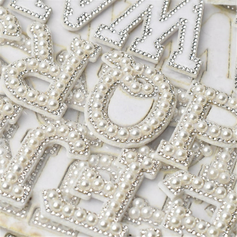 May include: A close-up of a collection of white felt letters decorated with pearl beads. The letters are arranged in a random order and are all the same size.
