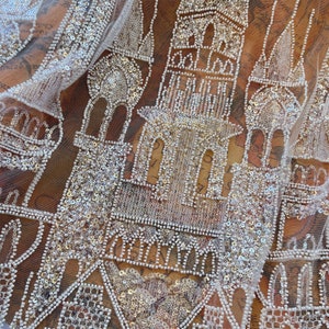 Può includere: Primo piano di un tessuto trasparente con un intricato disegno di elementi architettonici, tra cui torri e archi, creato con perline e paillettes bianche. Il disegno è posto su uno sfondo caldo e fantasia, creando un aspetto strutturato e dettagliato.