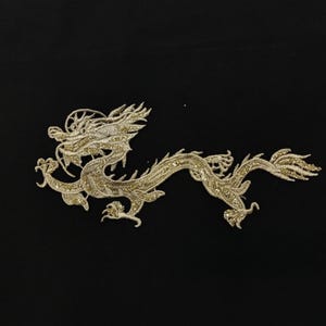 Può includere: Toppa a forma di drago ricamata in oro su sfondo nero. Il drago è finemente dettagliato con un corpo serpentino, piedi artigliati e una testa elaborata. Il ricamo presenta un filo d'oro scintillante.