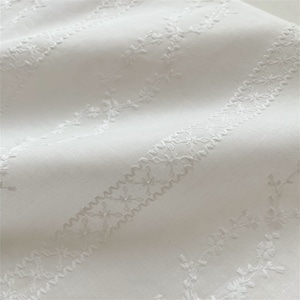 May include: White fabric with a delicate embroidered pattern of small flowers and leaves. The embroidery is in a repeating pattern of diagonal lines.