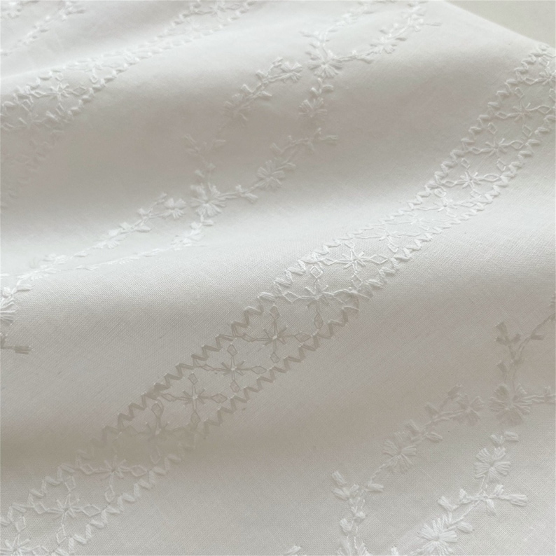 May include: White fabric with a delicate embroidered pattern of small flowers and leaves. The embroidery is in a repeating pattern of diagonal lines.