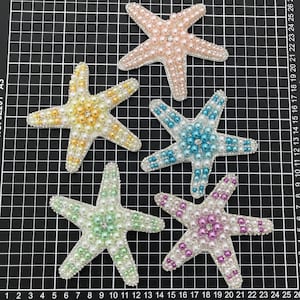 May include: Five starfish-shaped appliques arranged on a black grid mat. Each applique is covered in small white pearls and colored beads, including pink, yellow, blue, light green, and purple, with a single clear rhinestone at the center.