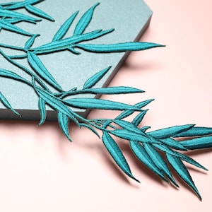 May include: Close-up of teal embroidered leaf sprigs. The leaves are long and slender, with detailed stitching. The sprigs are arranged on a grey surface against a pink background. The embroidery has a textured appearance.