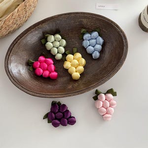 May include: Six fabric grape clusters in various colors including light green, blue, yellow, hot pink, purple, and light pink. Each cluster features small satin-like spheres arranged in a bunch with a dark green fabric leaf attached at the top.
