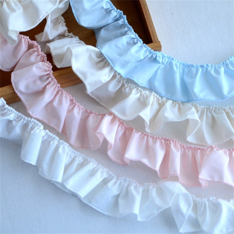 May include: Close-up of several rows of ruffled fabric trim in shades of white, light pink, and light blue. The fabric is gathered and sewn to create a decorative edge. The trim is ideal for sewing and craft projects.
