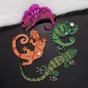 Könnte beinhalten: Vier bestickte Tier-Applikationen, darunter ein rosa Chamäleon, ein orangefarbener Gecko und zwei grüne Eidechsen. Die Applikationen sind mit bunten Perlen und Pailletten verziert.