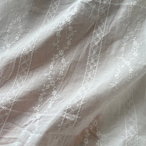 May include: White cotton fabric with delicate embroidered floral patterns in a repeating design. The fabric is sheer and has a soft, flowing texture.