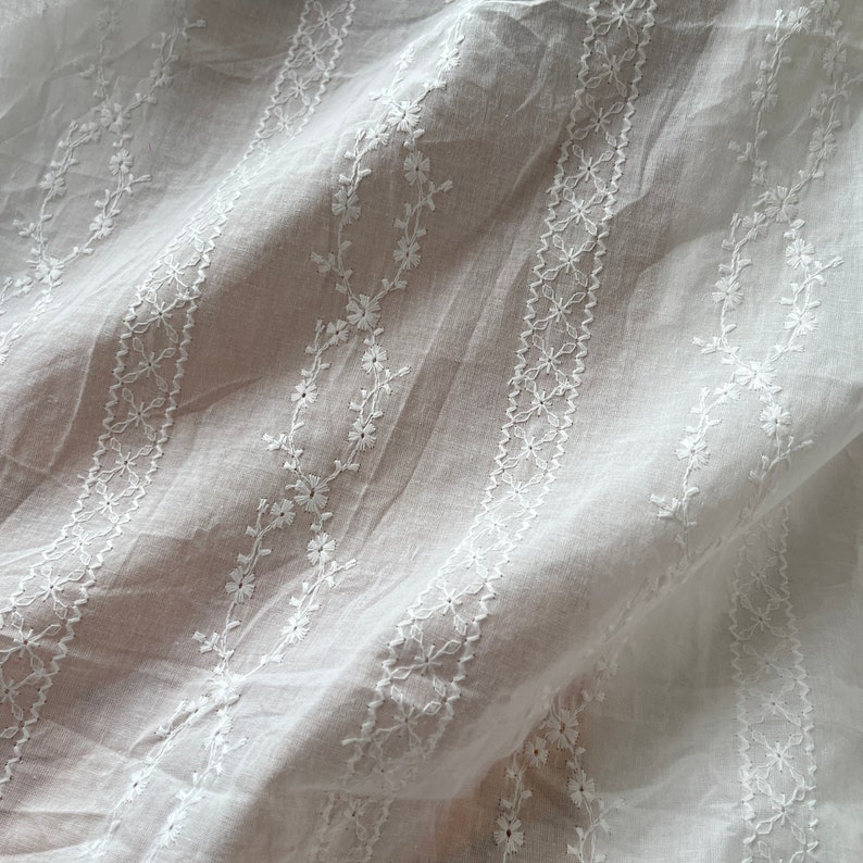 May include: White cotton fabric with delicate embroidered floral patterns in a repeating design. The fabric is sheer and has a soft, flowing texture.