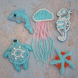 May include: A collection of beaded sea creature pins, including a dolphin, jellyfish, seahorse, turtle, and starfish. The pins are primarily turquoise, with accents of pink, white, and orange. Each pin is intricately detailed with small beads.