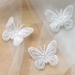 May include: Three white butterfly appliques crafted with intricate beadwork and sequins. Each butterfly features delicate antennae and detailed wing patterns, positioned on a sheer, translucent white fabric overlay with a light texture.