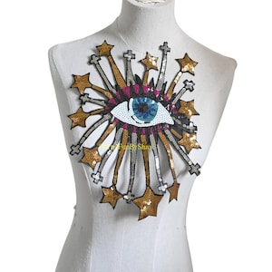 May include: A gold and silver sequined eye patch with a blue eye and pink eyelids. The eye is surrounded by gold stars and silver rays.