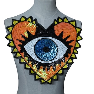May include: A heart-shaped sequin patch with a large blue eye in the centre. The heart is outlined in yellow and has a black outline around the eye.