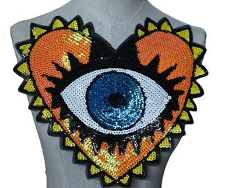 Könnte beinhalten: Ein herzförmiger Pailletten-Patch mit einem großen blauen Auge in der Mitte. Das Herz ist gelb umrandet und hat einen schwarzen Rahmen um das Auge.