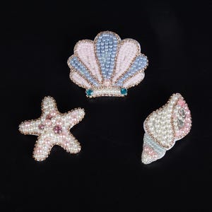 Peut inclure: Trois écussons décoratifs en forme de coquille Saint-Jacques, d'étoile de mer et de conque. Chaque pièce est minutieusement ornée de petites perles blanches, roses et bleu clair, ainsi que de cristaux transparents, bordés de fil doré.