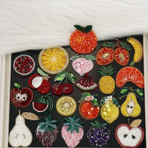 Può includere: Una collezione di toppe a forma di frutta realizzate con perline e paillettes colorate. Il set include rappresentazioni di arance, limoni, fragole, ciliegie, pere, mele, ananas, fette di anguria e una banana, tutte caratterizzate da intricate perline.