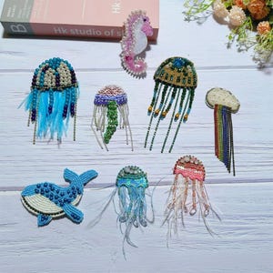May include: A collection of seven beaded and embroidered sea creature patches, including a pink seahorse, a blue whale, and several jellyfish in various colours. The patches are all handmade and feature intricate details.
