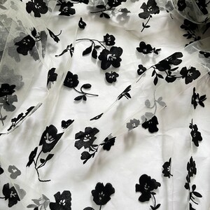 May include: White sheer fabric with black floral embroidery. The fabric is delicate and has a soft, flowing texture.