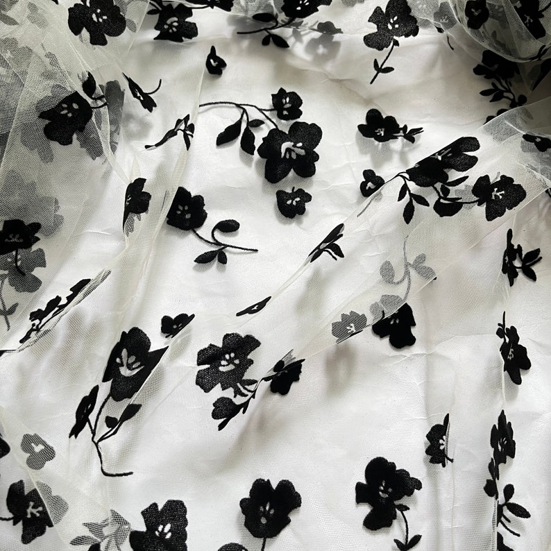 May include: White sheer fabric with black floral embroidery. The fabric is delicate and has a soft, flowing texture.