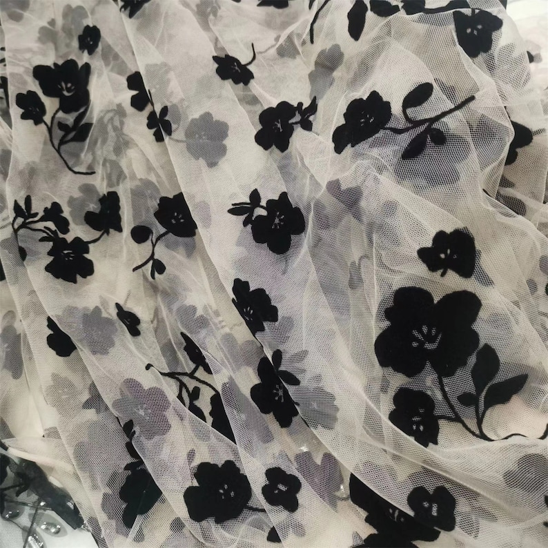 May include: White sheer fabric with black velvet floral embroidery. The fabric is delicate and has a soft, flowing texture.