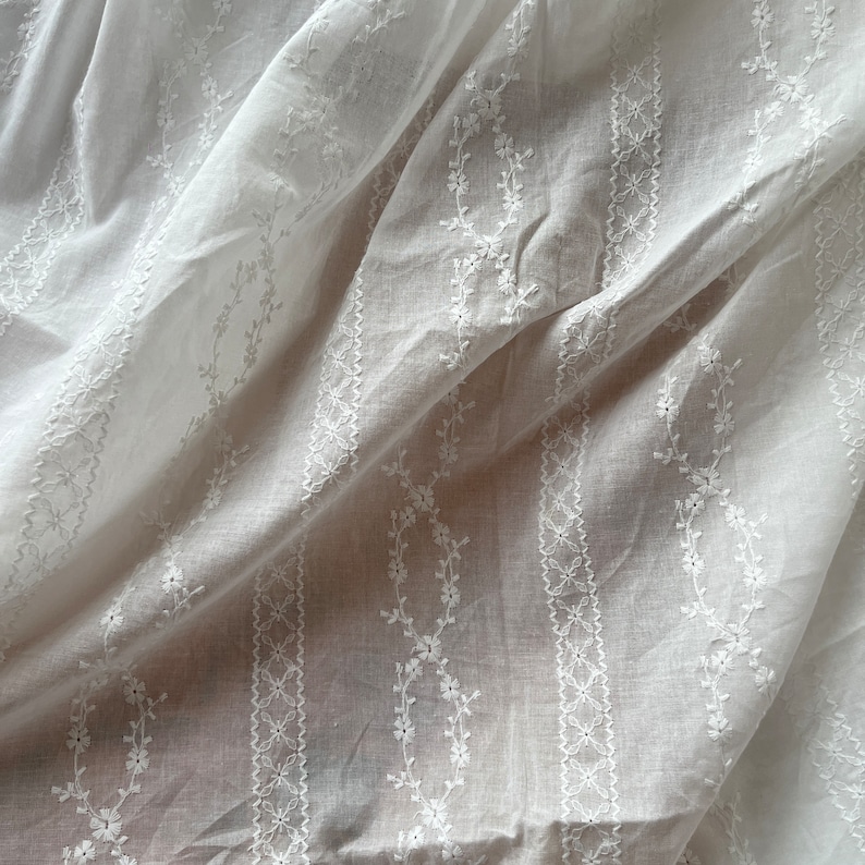 May include: White cotton fabric with delicate embroidered floral patterns in vertical stripes. The fabric is sheer and has a soft, flowing texture.