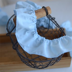 May include: A light blue fabric ruffle adorns the rim of a vintage wire basket. The basket has a wooden handle and a dark metal frame. The fabric is gathered and sewn, creating a delicate, ruffled edge. The basket sits on a wooden surface.