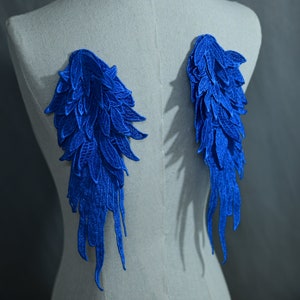Pode incluir: Duas asas de renda azul com um design de penas. As asas estão presas a um fundo de tecido branco.