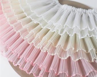 Può includere: Una gonna a più strati caratterizzata da una sfumatura di colori che passa dal bianco in alto al giallo pallido, verde chiaro e rosa tenue in basso. La gonna è realizzata in tessuto plissettato e trasparente con delicati bordi smerlati.