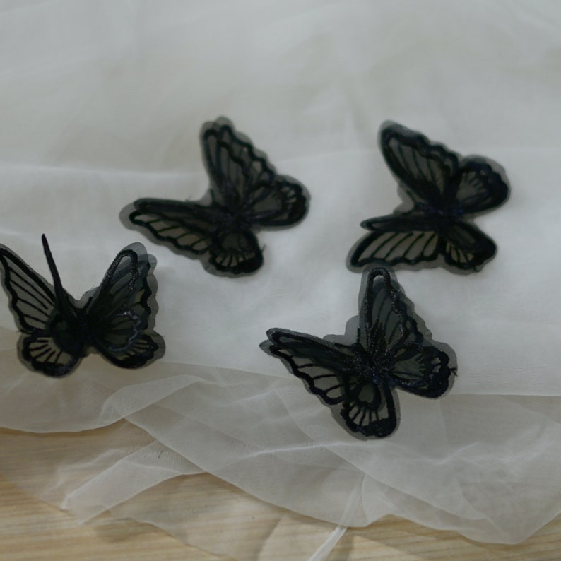 May include: Four black butterfly embellishments with detailed wings are displayed on a white, translucent fabric. The butterflies have a delicate, lacy appearance, suggesting they might be used for crafts or decorative purposes.