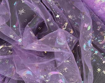 Pode incluir: Close-up de um tecido de tule lavanda com um design celestial. O tecido é adornado com estrelas e luas crescentes iridescentes. O tule é reunido e drapeado, criando um efeito macio e fluido. Ideal para artesanato, fantasias ou decoração.