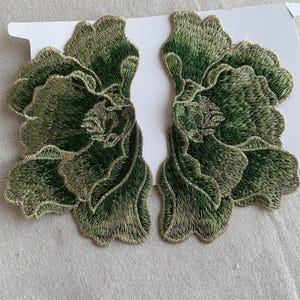May include: Two embroidered floral appliques. Each applique features a detailed design of a flower with layered petals. The embroidery uses shades of green and gold thread, creating a textured and dimensional effect. The appliques are on a white backing.