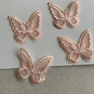 May include: Four light pink butterfly appliques. Each butterfly has detailed embroidery and sheer, translucent wings. The appliques are set against a white background, highlighting their intricate design and craftsmanship.