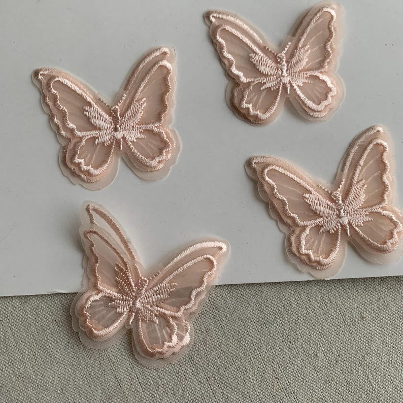 May include: Four light pink butterfly appliques. Each butterfly has detailed embroidery and sheer, translucent wings. The appliques are set against a white background, highlighting their intricate design and craftsmanship.