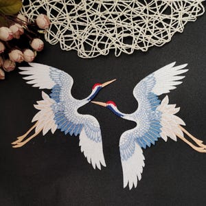 May include: Two white embroidered crane patches with blue and pink accents. The cranes are facing each other with their wings spread out.