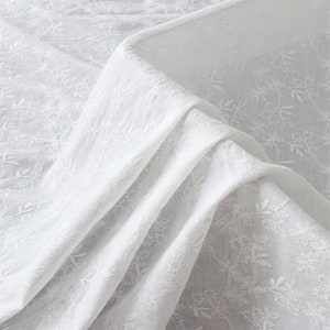May include: White cotton fabric with a delicate floral embroidery pattern. The fabric is folded and draped, showcasing the intricate details of the design.