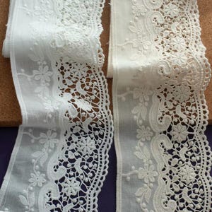Op de afbeelding: Twee stroken witte kanten bies met een ingewikkeld geborduurd bloemenpatroon. Het kant heeft een geschulpte rand en een transparante, lichtgewicht stoffen basis met gedetailleerde borduurmotieven van bloemen, bladeren en krullende ranken.
