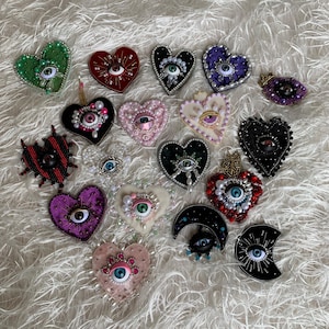 Peut inclure: Une collection de broches en forme de cœur et de croissant de lune avec des motifs d'yeux centraux. Chaque pièce est ornée de perles, de paillettes et de perles dans diverses couleurs, notamment le rouge, le vert, le noir, le violet, le rose et le blanc.