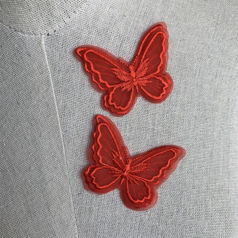 May include: Two red butterfly patches with detailed embroidery. The patches are made of a sheer material, and the edges are outlined with a darker red thread. The butterflies are displayed on a light grey fabric.